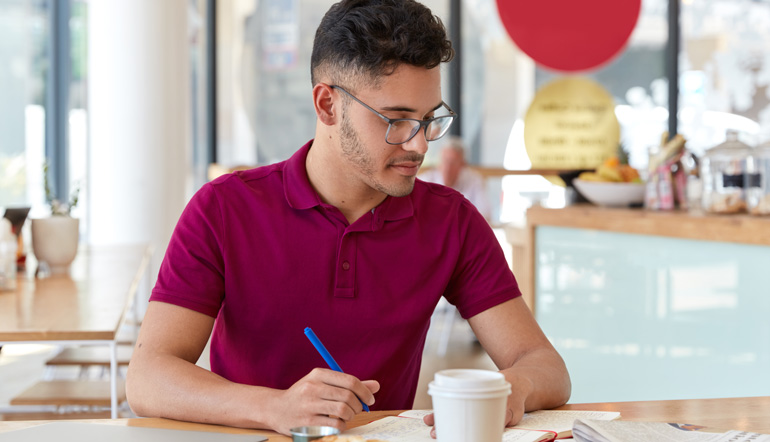 Photo of mixed race male student writes necessary information in notepad from daily newspaper, creates simmilar article