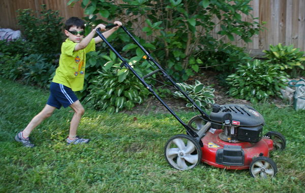 young boy with a green t-shirt, blue shorts, and sunglasses pushing a lawnmower